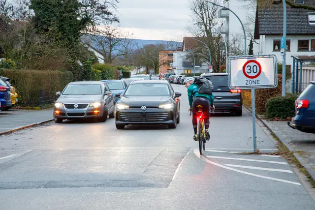 Ein Fahrzeug im Halteverbot und ein weiteres Auto behindern den Verkehr. Radfahrer und querende Schülerinnen und Schüler teilen sich den verbleibenden Raum.